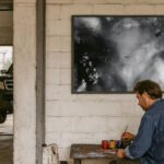 Künstler beim Malen vor weißer Backsteinwand – abstraktes Schwarz-Weiß-Gemälde über Werkbank in industrieller Umgebung