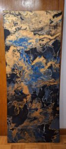 Abstract acrylic pour painting in black, gold and cobalt blue, 150 x 61 cm. The canvas narrows slightly in the middle, giving it an organic, non-rectangular shape. Within the layered pigments, hidden forms emerge depending on orientation: small animals, a folded brain, and a grim face visible when the painting is turned horizontally. Created by emerging artist Manuela Gawehn.